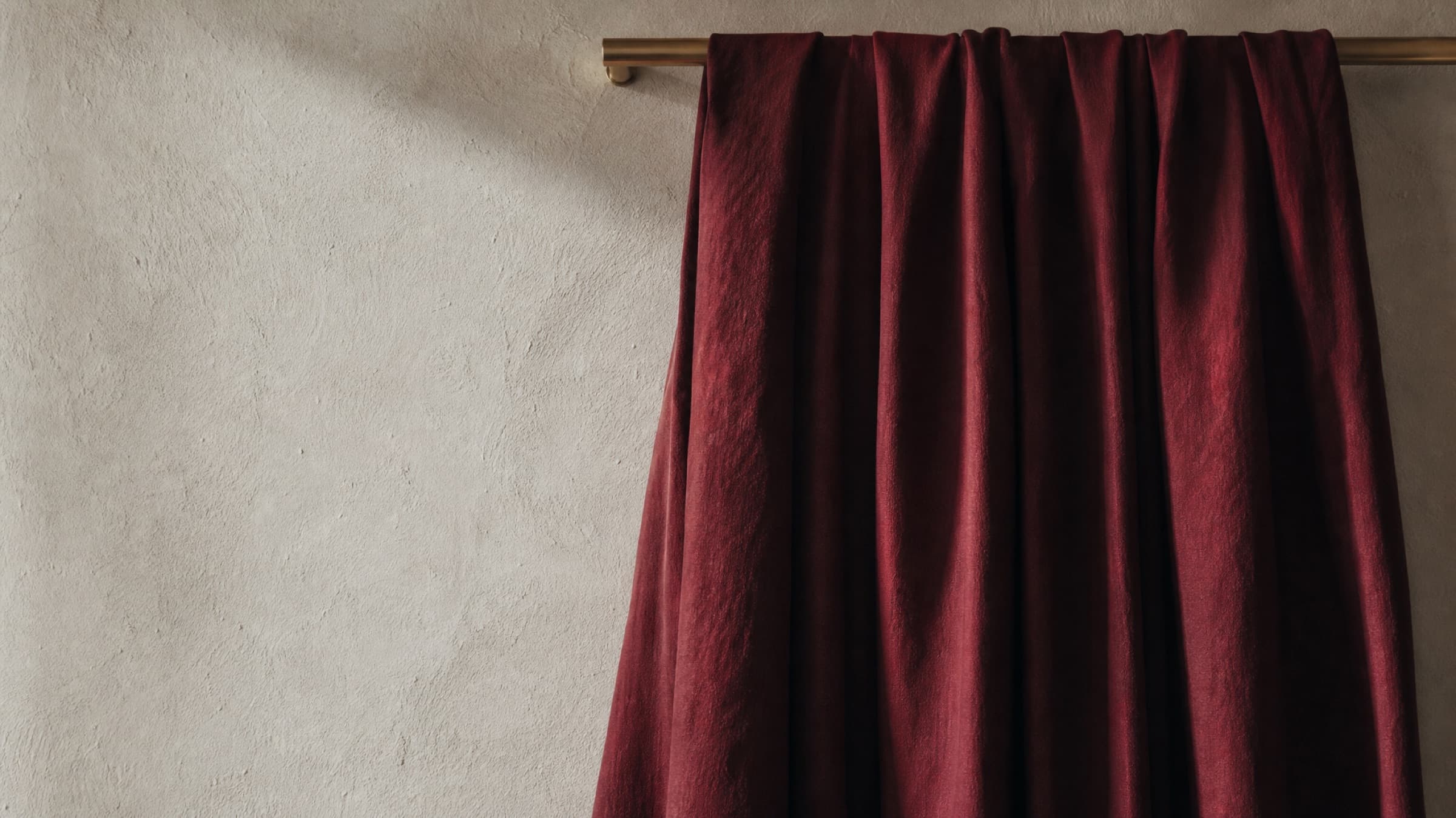 A length of deep wine-red cloth hung from a brass rail, falling in deep vertical folds against a limestone plaster wall.