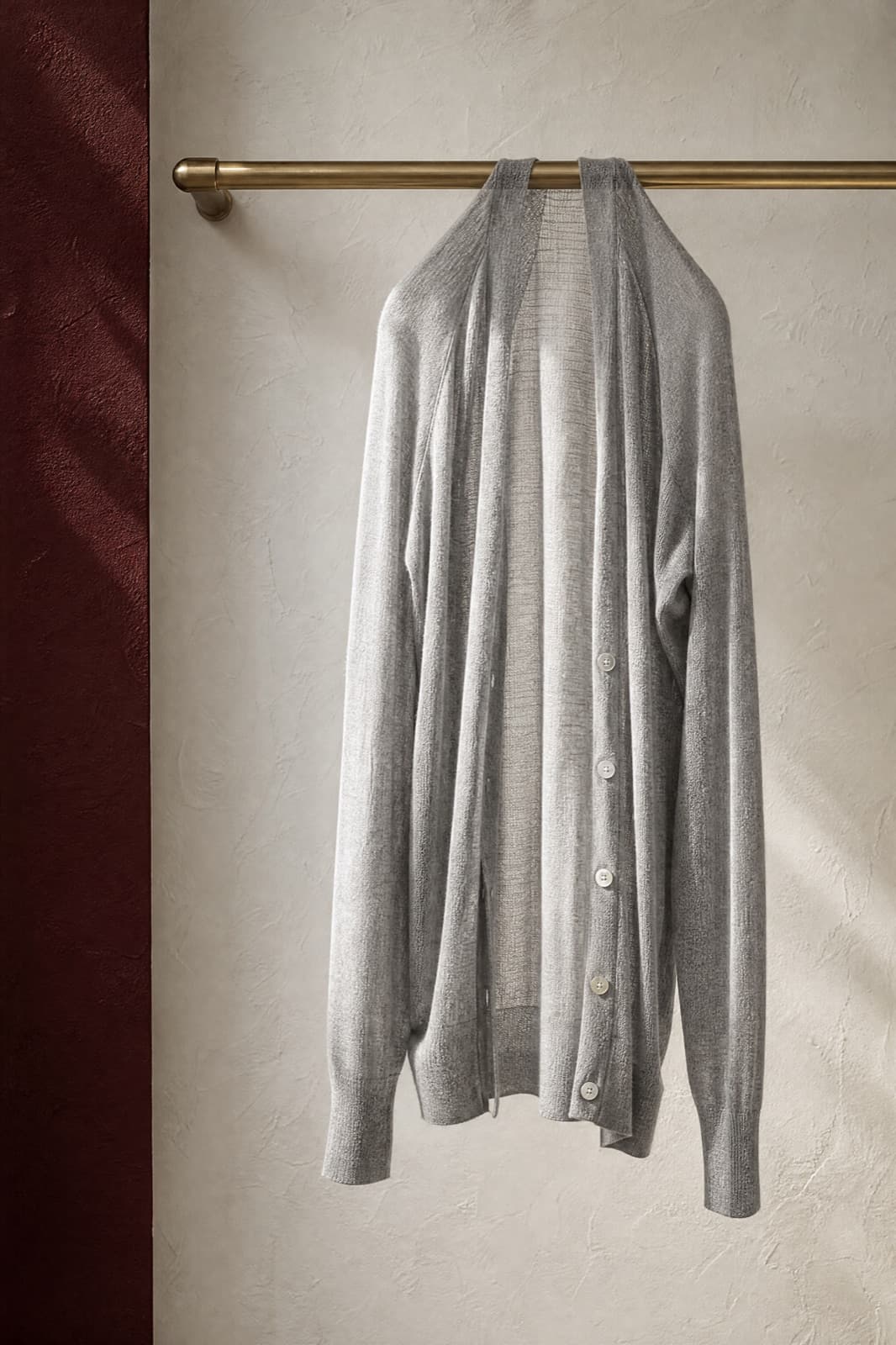 Fine-Knit Cardigan, hung from a rail against limestone plaster.