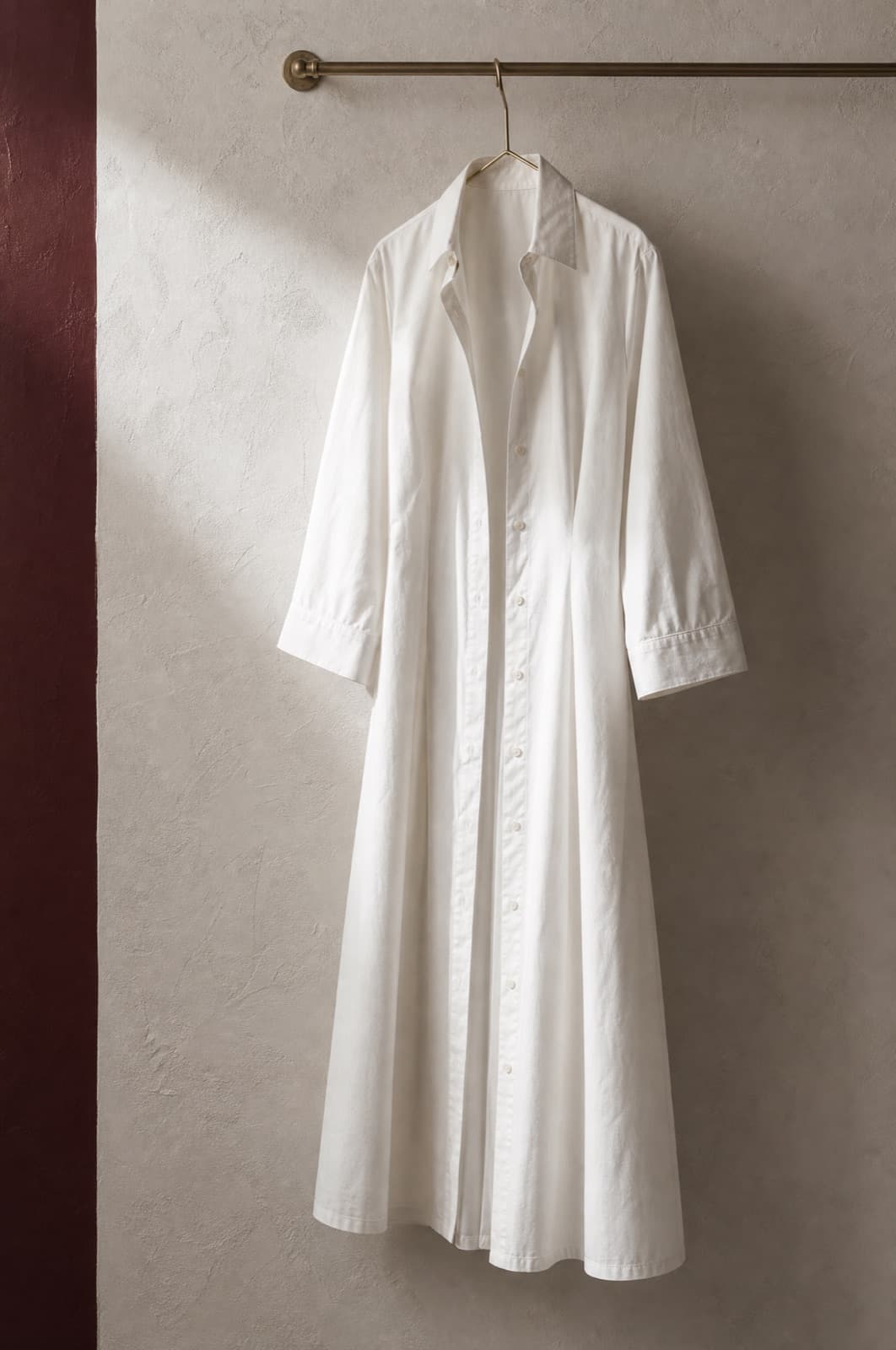 Sedaine Shirt Dress Midi, hung from a rail against limestone plaster.